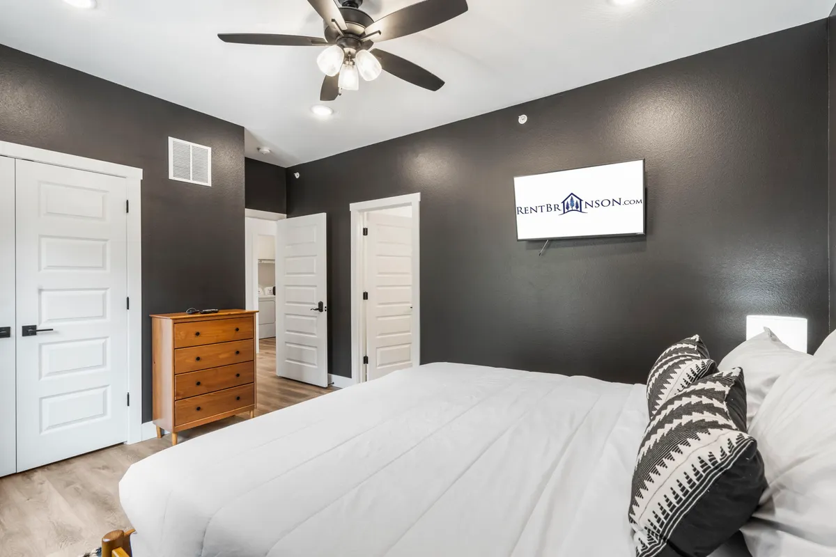 A tranquil, contemporary bedroom complete with a comfortable bed, ceiling fan, dresser, and a wall-mounted television. The decor is modern and inviting, with soft lighting that enhances the restful ambiance.