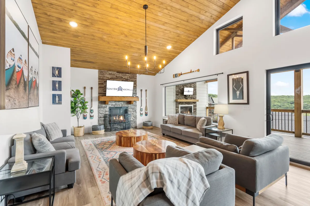 The image showcases a cozy living room with plush sofas gathered around a crackling fireplace. Expansive windows frame stunning lake views, filling the space with natural light and offering a perfect spot to unwind and watch the sunset. The inviting setup suggests a space ideal for relaxing, socializing, or enjoying a warm drink by the fire.