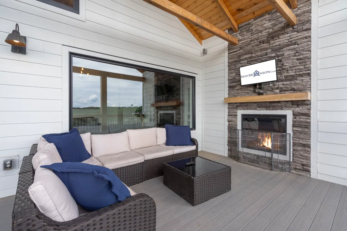 A stylish outdoor seating area featuring a large, stone fireplace, cozy sofas, and a mounted television, with expansive windows framing the picturesque natural surroundings. The space is designed for socializing, relaxation, and embracing the outdoors, day or night.
