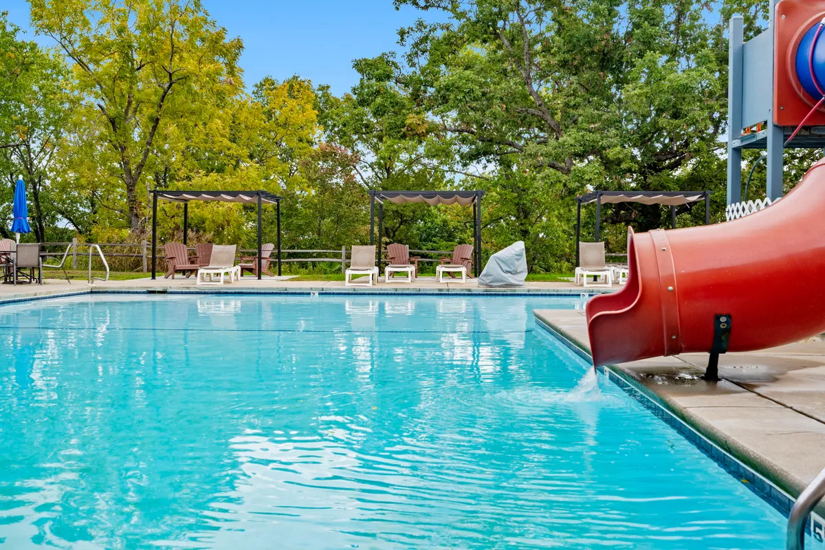 A bright, playful pool scene featuring a large pool with a water slide, lounge chairs, canopies, and surrounding trees. The area is filled with the energy of children playing and families gathering, highlighting the resort's appeal for group activities and summer fun.