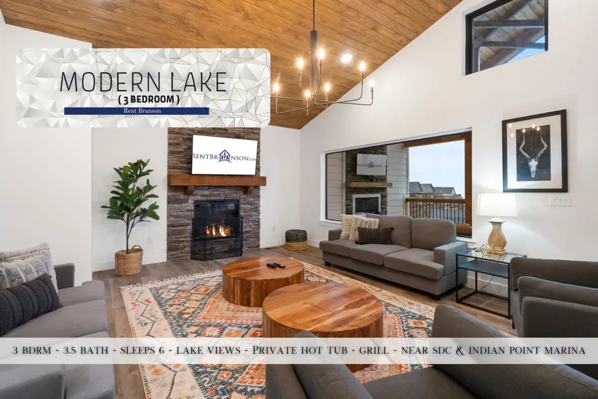A welcoming living room with a plush sofa arrangement, a glowing fireplace, coffee table, decorative artwork, and expansive windows looking out over the lake. The space exudes warmth and togetherness, ideal for family gatherings and cozy evenings.