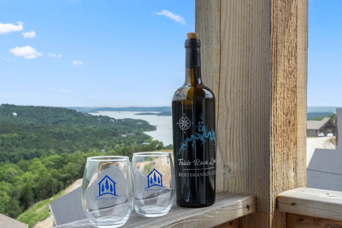 A serene exterior deck scene featuring a bottle of wine and two glasses set on a table, overlooking the expansive, scenic view of Table Rock Lake. Gentle sunlight highlights the calm water and distant hills, evoking a sense of tranquility and luxury.