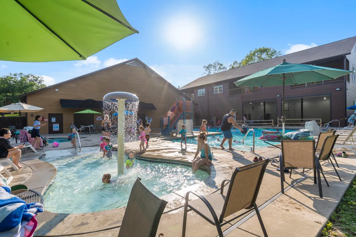 A lively pool area where children play in the water fountain, adults relax under sun umbrellas, and the atmosphere is bright and cheerful. The scene is full of social energy, emphasizing the family-friendly and active elements of the neighborhood.