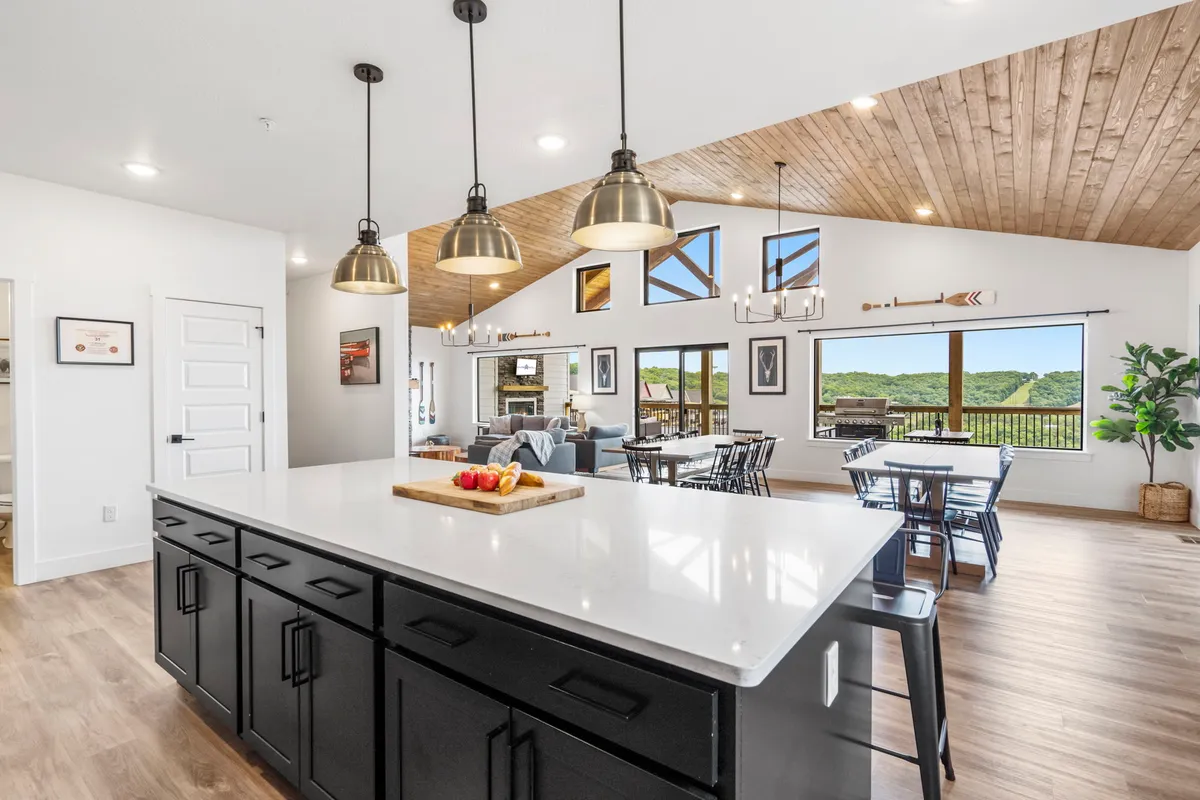 This image showcases a bright, open kitchen with a spacious island countertop, pendant lighting, and large windows that offer views of the landscape. The kitchen seamlessly flows into a dining area, emphasizing the open-concept design.